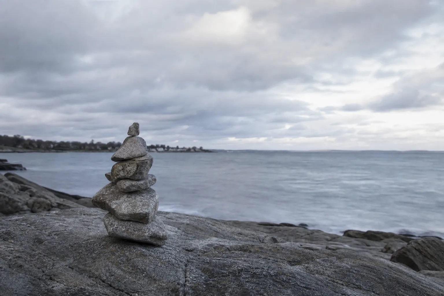 A small stack of balanced stones on a rocky shoreline beneath heavy clouds, with calm gray water stretching to the horizon.