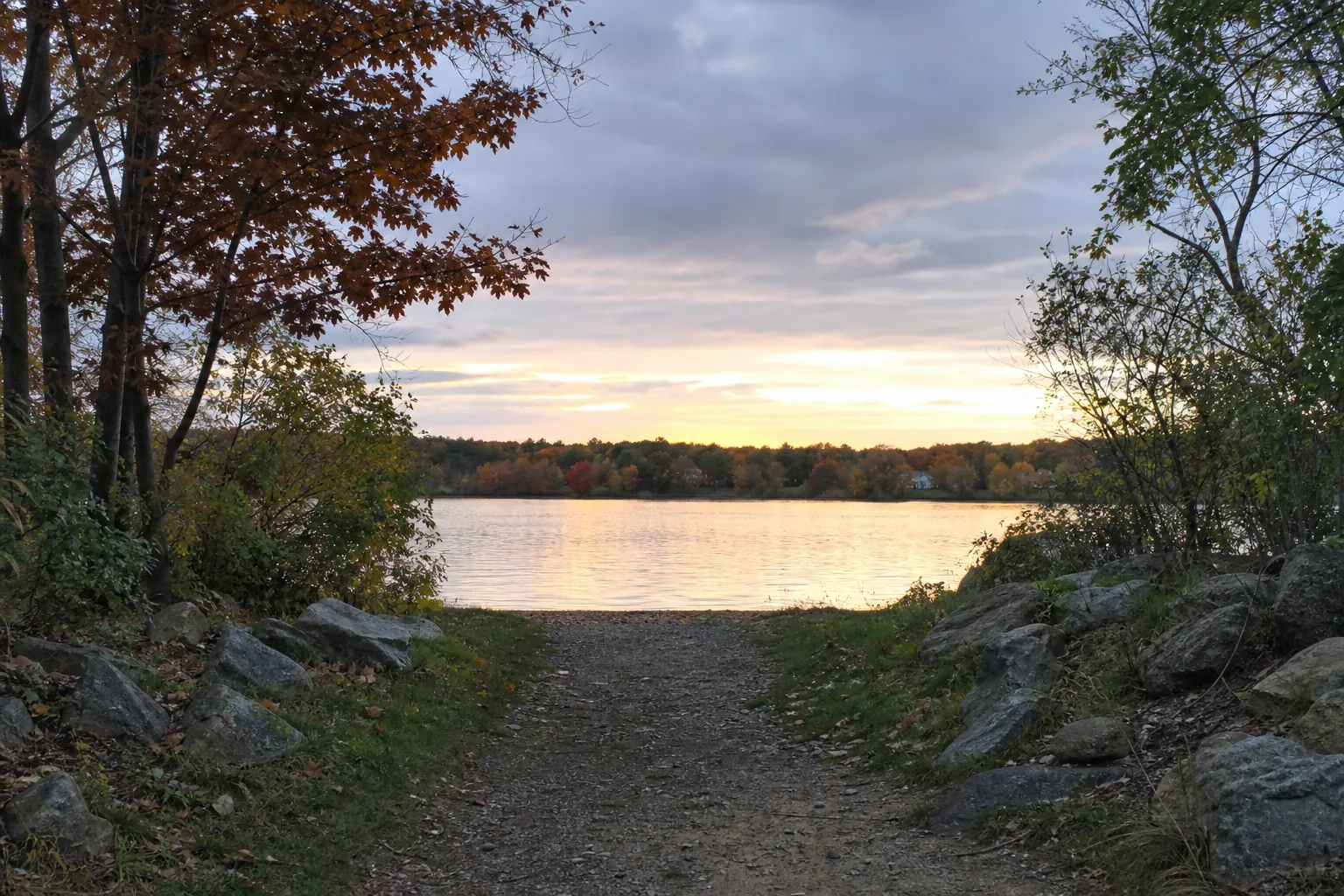 Autumn path leading to a lakeside sunset A rocky dirt path leads through autumn trees to a calm lake beneath a cloudy sunset sky.