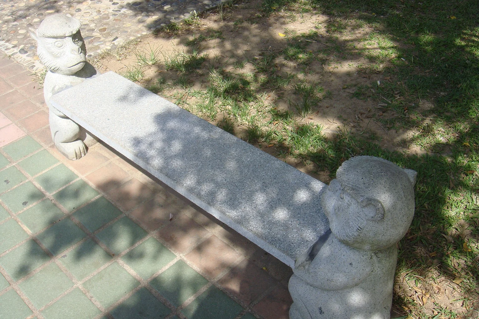 Stone bench with carved supports beside patterned paving A pale stone bench with carved supports stands beside patterned paving and grass in dappled shade.