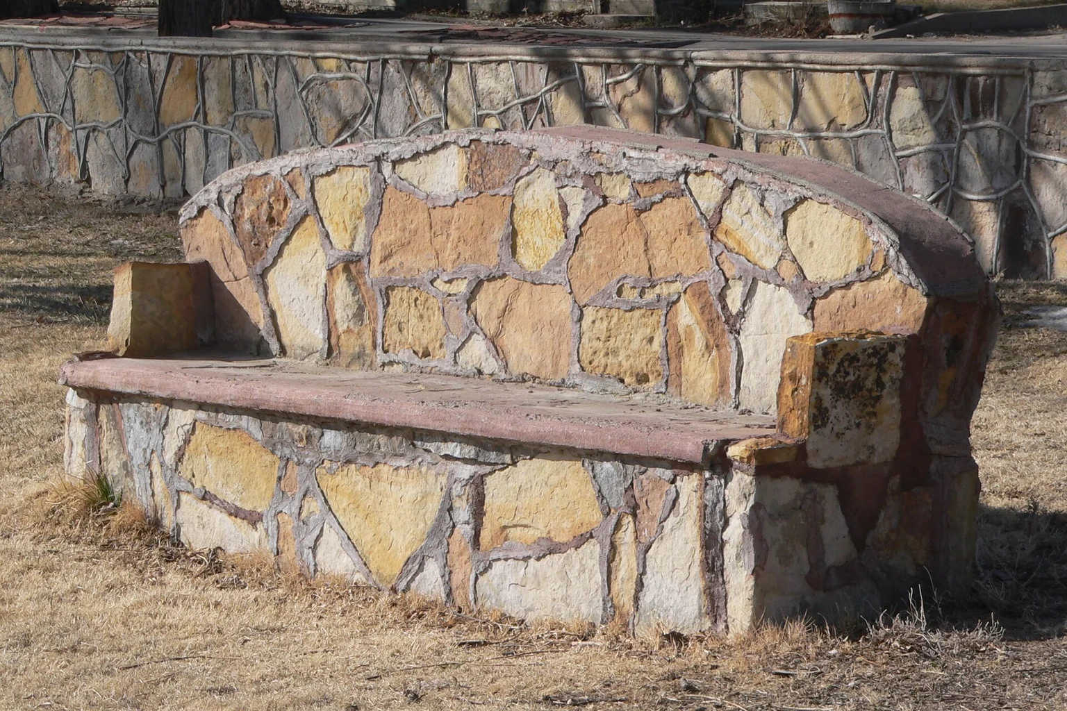 Curved stone bench on dry grass An empty curved stone bench sits on dry grass in front of a matching stone wall.