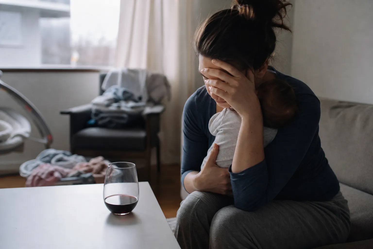 Distressed mother holding an infant on a couch beside a glass of red wine in a cluttered living room