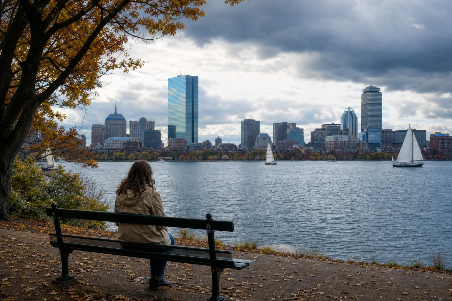A solitary person sits on a park bench across the Charles River from the Boston skyline beneath dark autumn clouds.