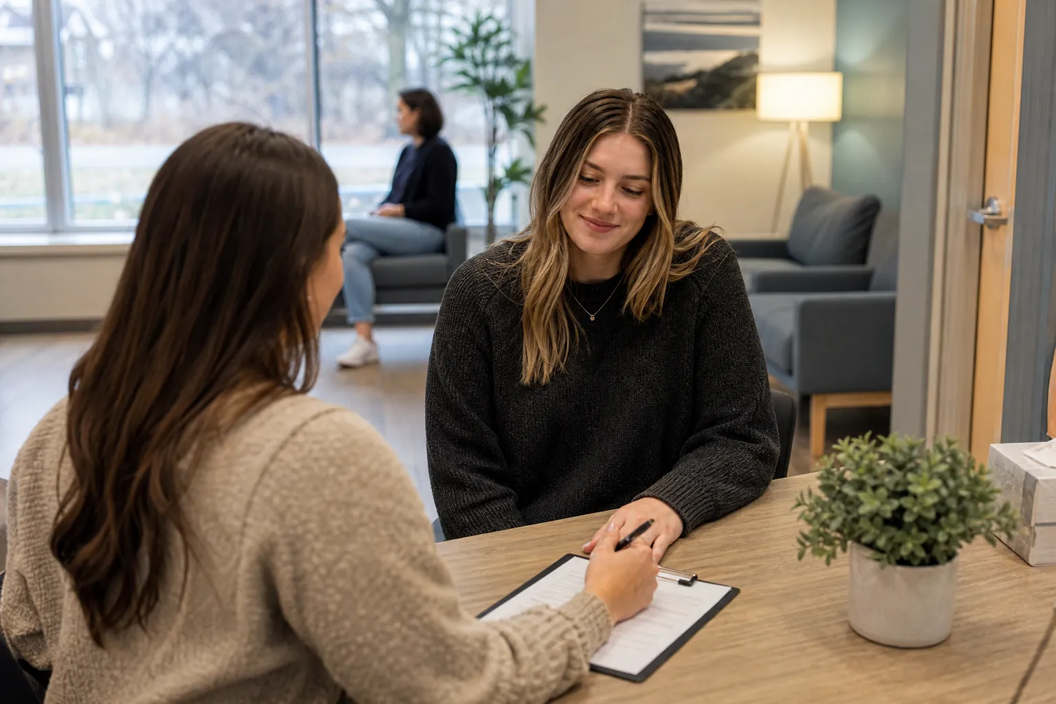 Arrival check-in at a mental health clinic reception desk A woman checks in with a staff member holding a clipboard at a reception desk in a comfortable clinic waiting room.