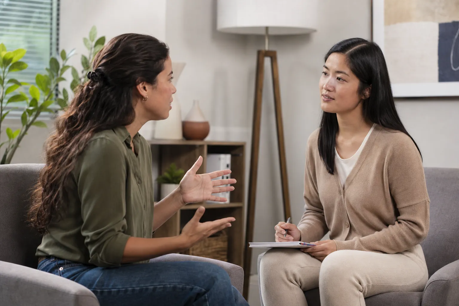 Two women sit facing each other in an office, one gesturing while the other listens with a pen and clipboard.