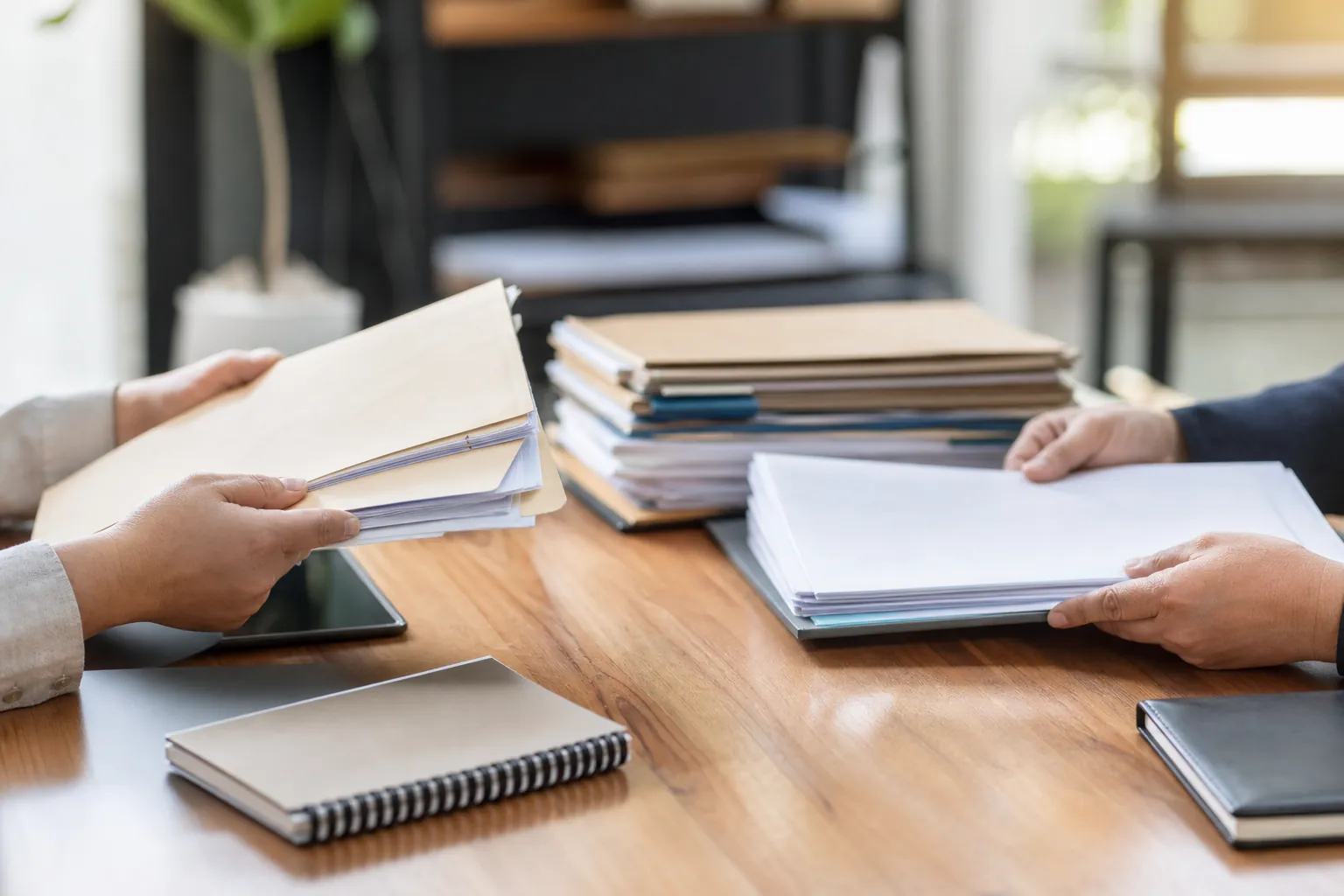 Two people hold folders and loose paperwork across a wooden table with stacked files, notebooks, and a tablet.