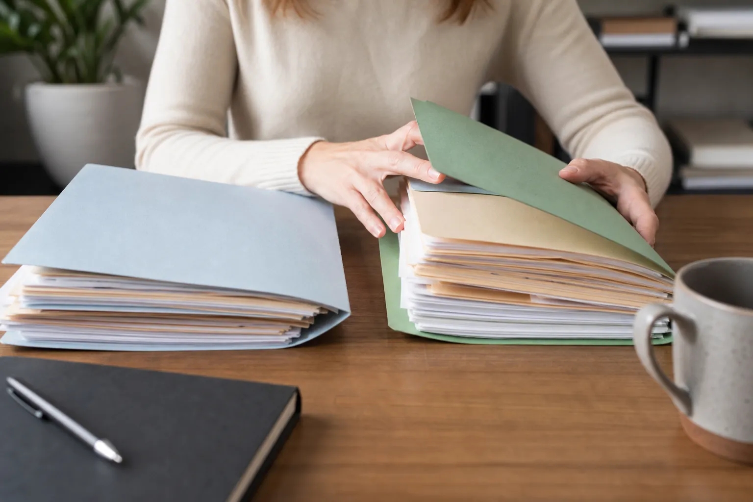 Sorting Papers in Separate Record Folders A person handles a green folder of papers beside a separate blue folder, a notebook, pen, and mug on a wooden desk.