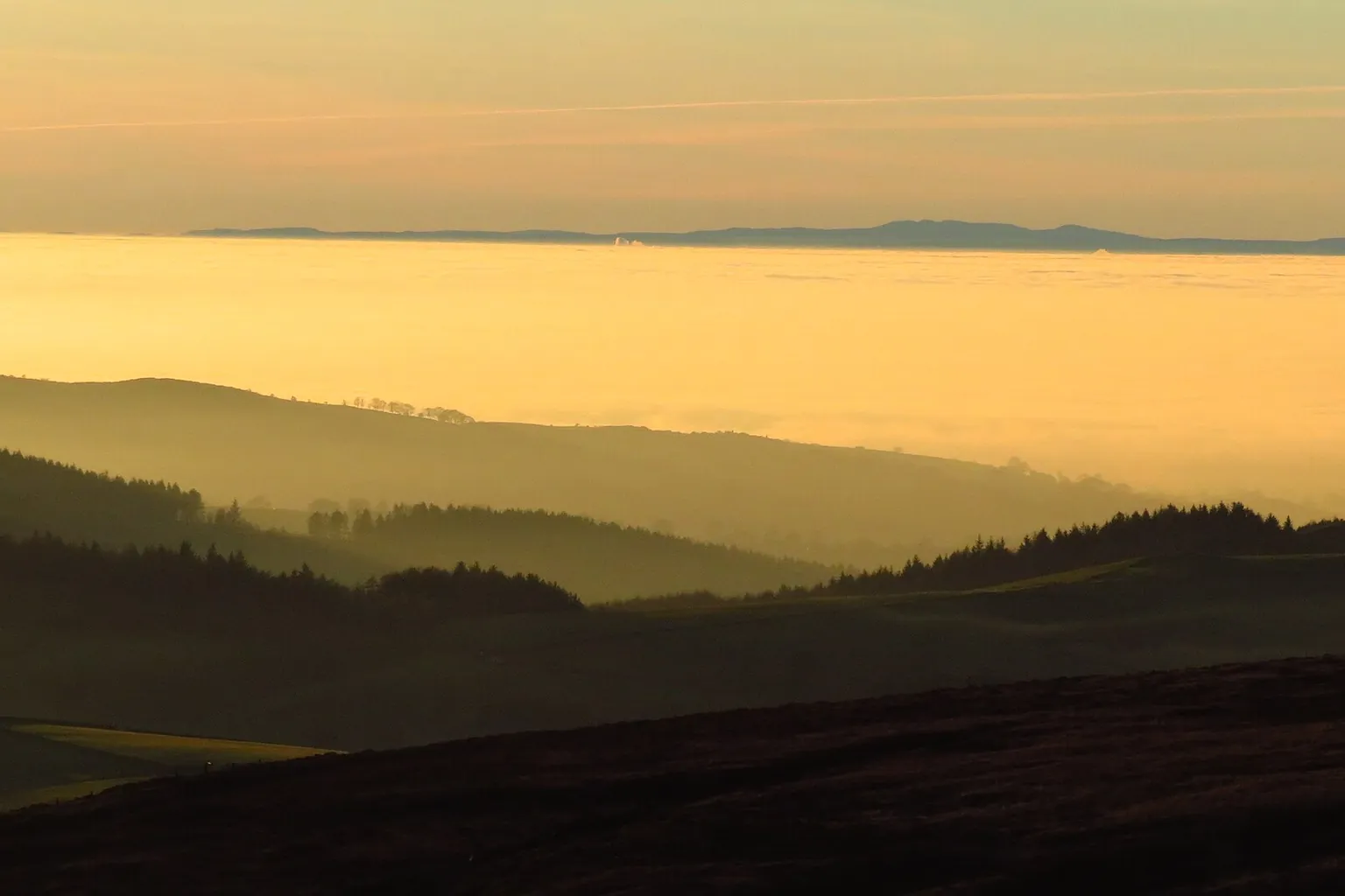 Layered hills and a dark ridge sit below a hazy orange sky, with low sunlight over the distant landscape.