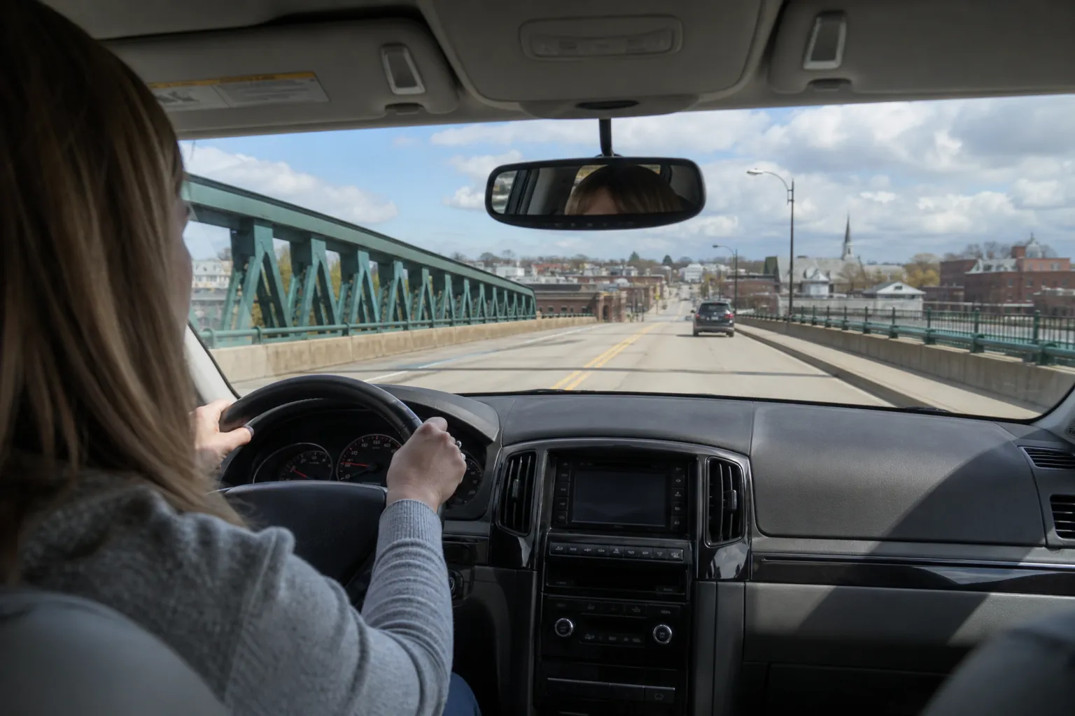 A driver holds the steering wheel while crossing a bridge toward town, viewed from the back seat of a car.