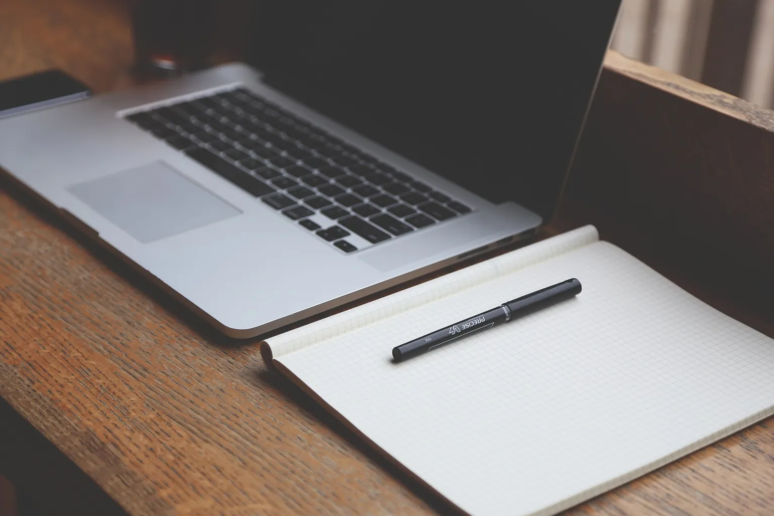 A laptop with a dark screen sits beside a blank notebook and pen on a warm wooden A laptop with a dark screen sits beside a blank notebook and pen on a warm wooden table.