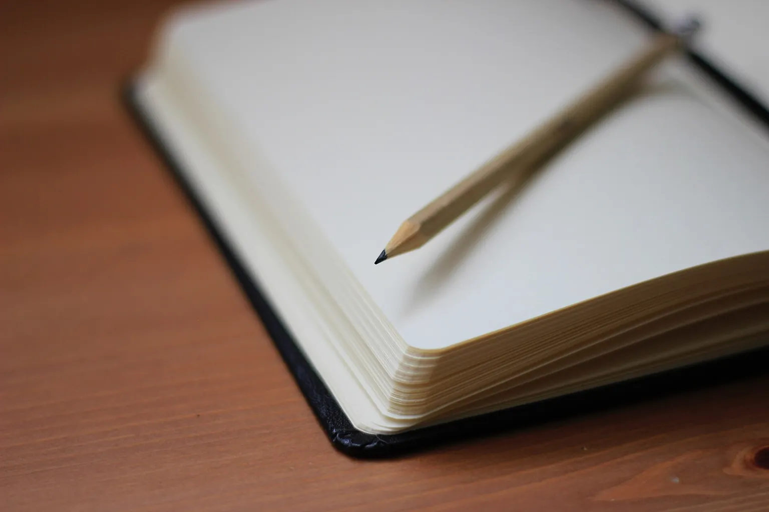 A pencil rests across the blank pages of an open notebook on a wooden surface in A pencil rests across the blank pages of an open notebook on a wooden surface in soft light.