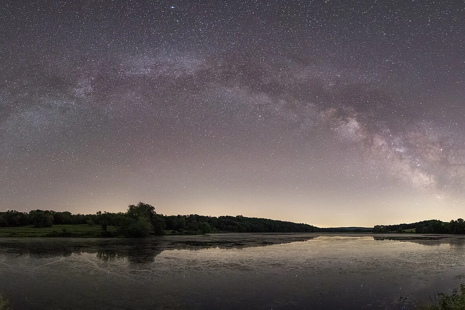 Stars and a faint Milky Way stretch over a still lake and a dark tree-lined shore.