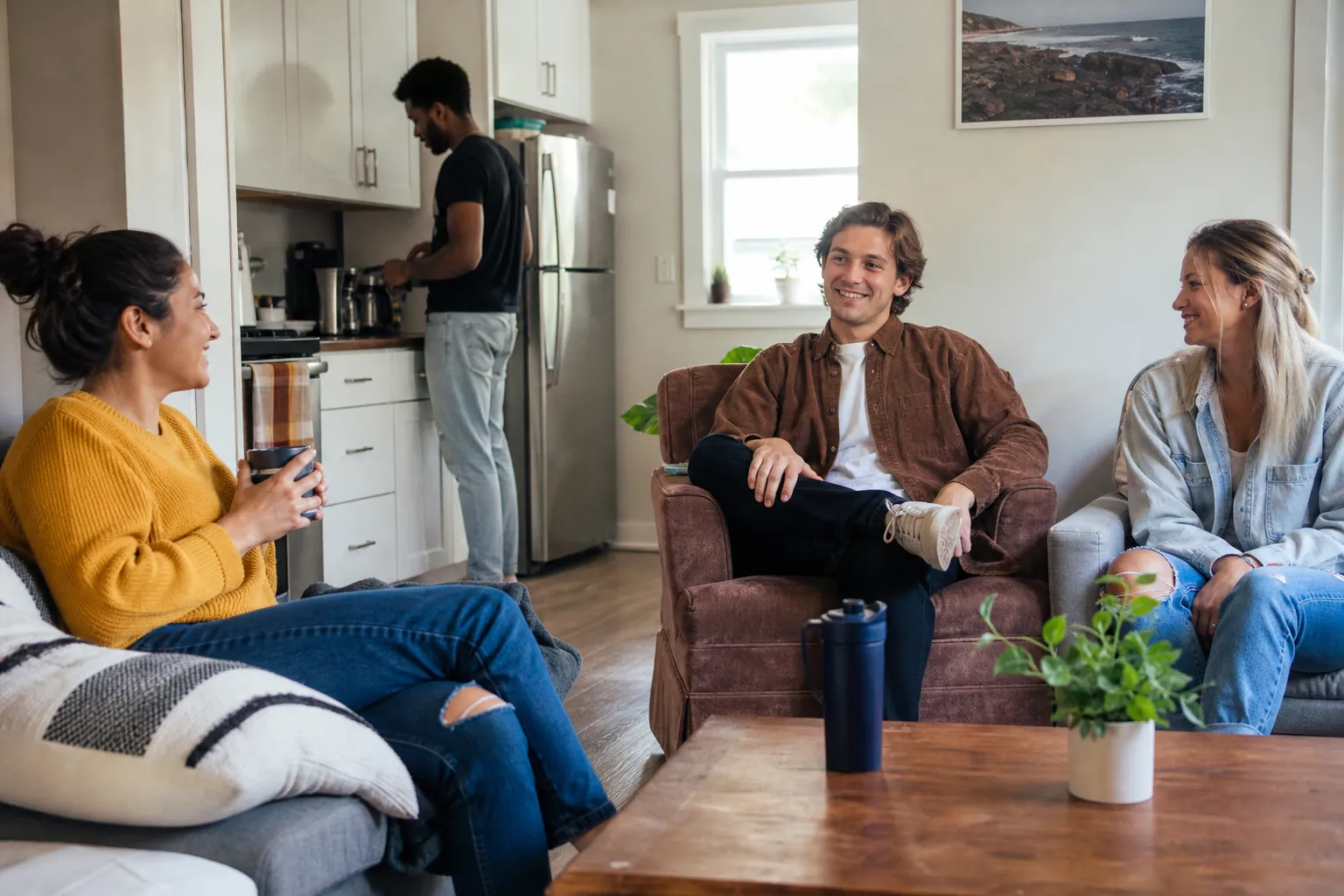 Adults Socializing in a Shared Living Room and Kitchen Three adults sit together in a living room while a fourth stands at the counter in the adjoining kitchen.