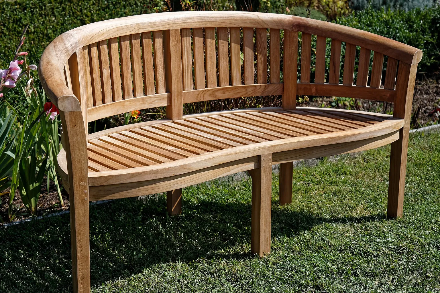 A curved wooden bench with a slatted seat stands in a sunlit garden beside grass and A curved wooden bench with a slatted seat stands in a sunlit garden beside grass and flowering plants.