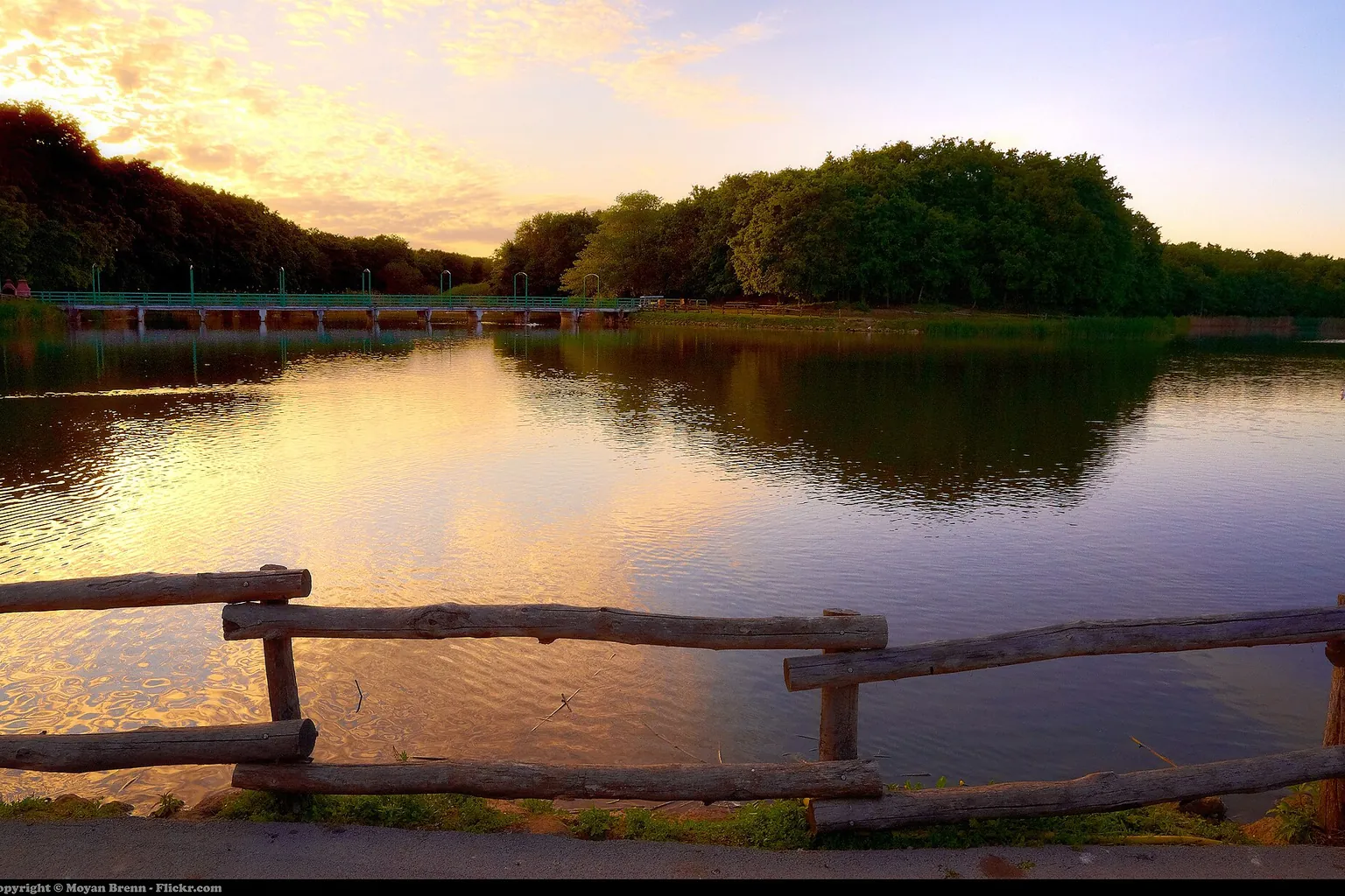 A wooden railing borders a still lake at sunset, with trees and a warm clouded sky reflected in the water.
