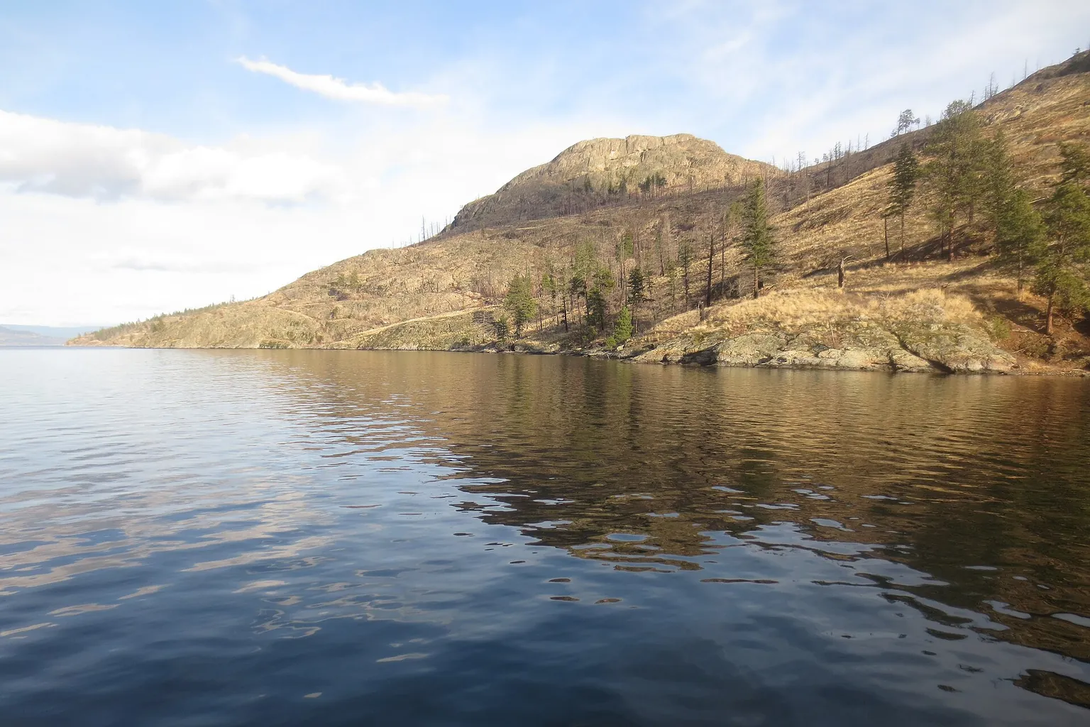 A calm lake lies beneath a rocky wooded hillside, with soft clouds and light ripples on the water.