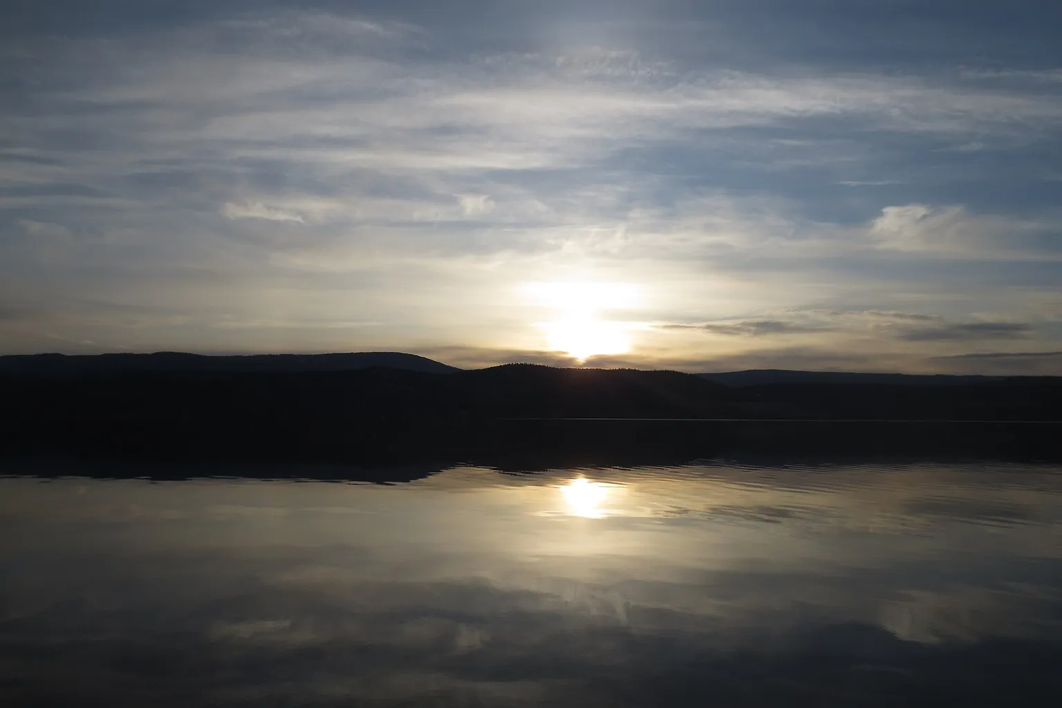 Low sunlight shines through clouds over a broad lake, with dark hills reflected in the still water.