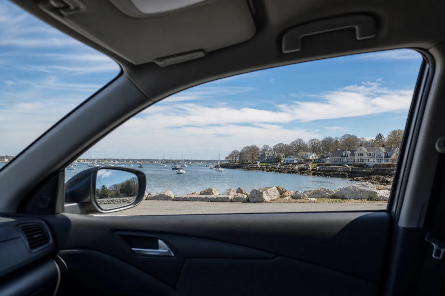 View through a car's passenger window of a coastal harbor with moored boats, shoreline rocks, and waterfront houses.