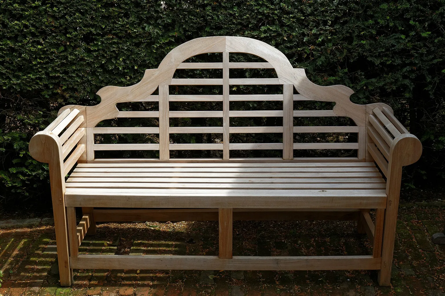 A pale wooden garden bench with a decorative curved back rests in front of dense green foliage.