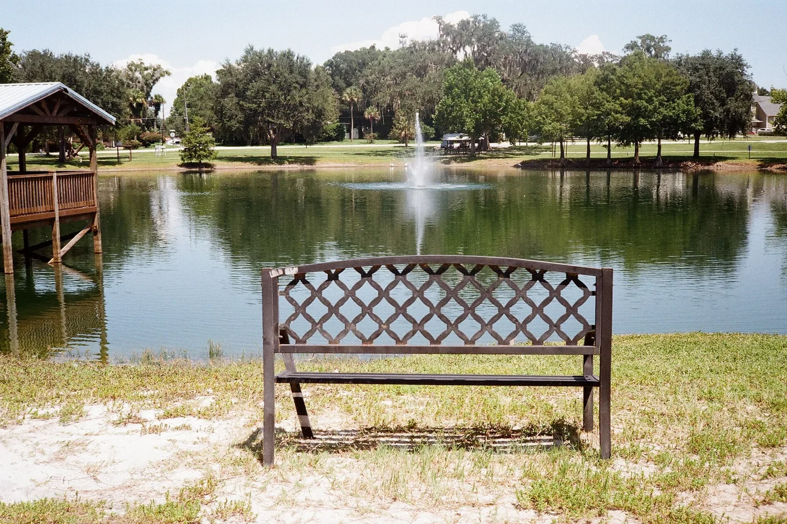 Bench beside a pond and fountain Empty metal bench beside a pond with a fountain, trees, and a wooden pavilion.