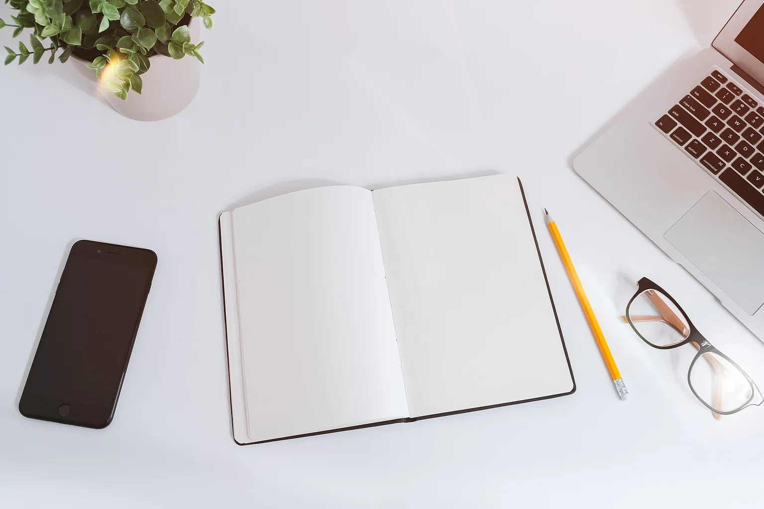 A blank open notebook lies on a white desk beside a dark phone, glasses, pencils, a A blank open notebook lies on a white desk beside a dark phone, glasses, pencils, a plant, and a laptop.