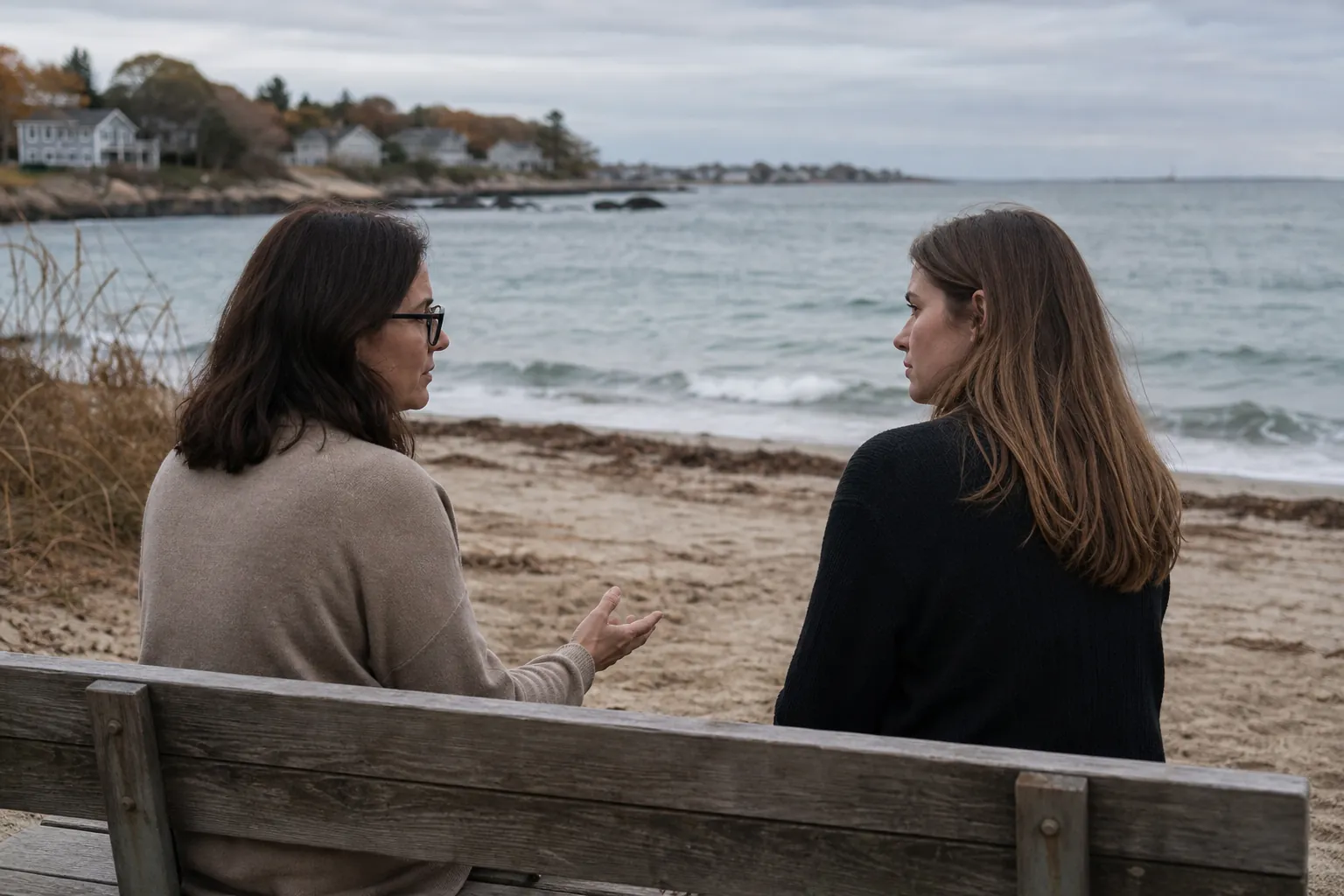 Two women sit facing each other on a wooden bench beside a beach, with one gesturing during their conversation.