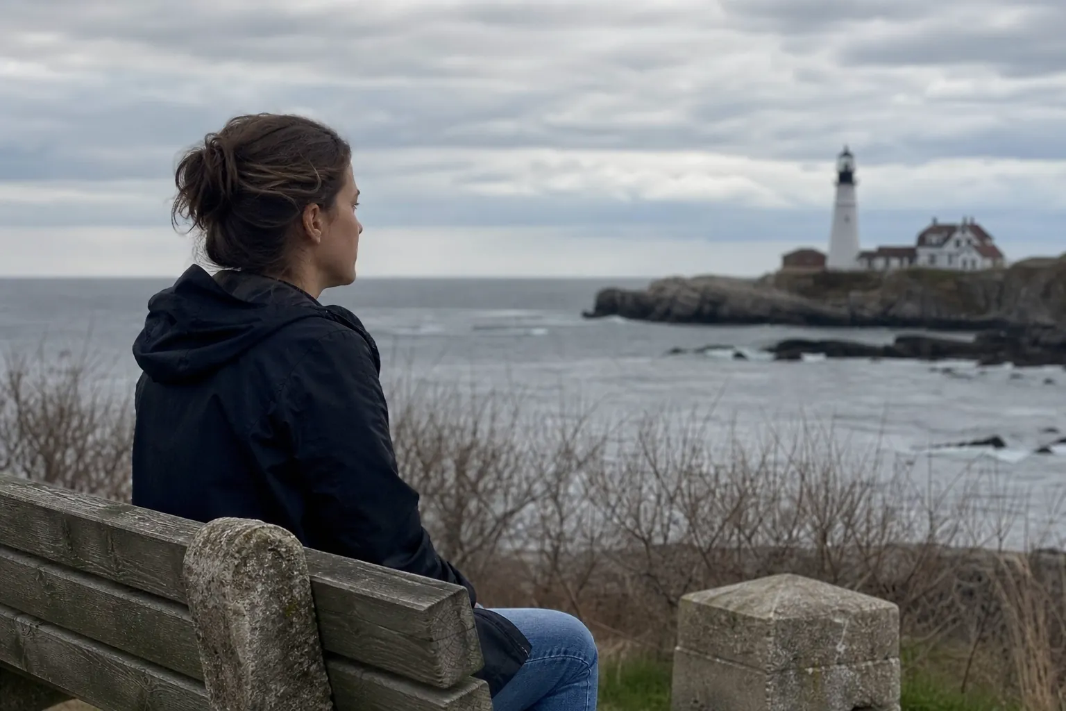 Woman Reflecting by the Ocean Near a Lighthouse A woman sits alone on a wooden bench, looking across a gray ocean toward a distant lighthouse under cloudy skies.