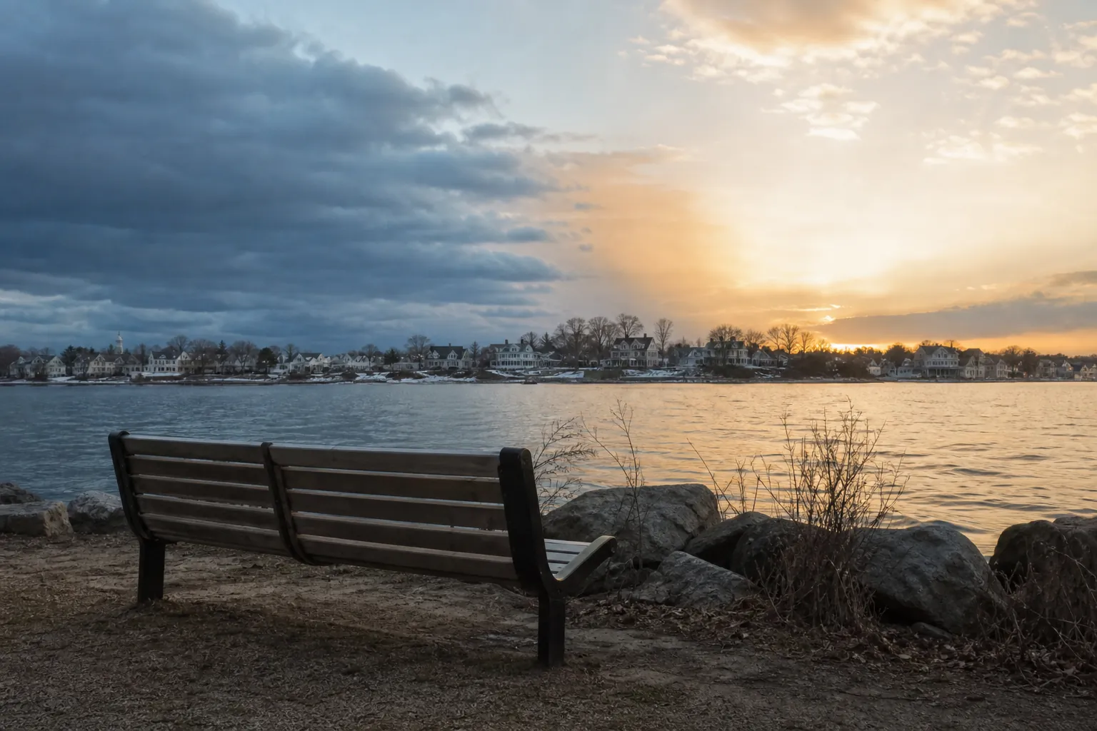 Empty wooden bench facing calm water and a coastal town beneath dark clouds and a warm sunset.