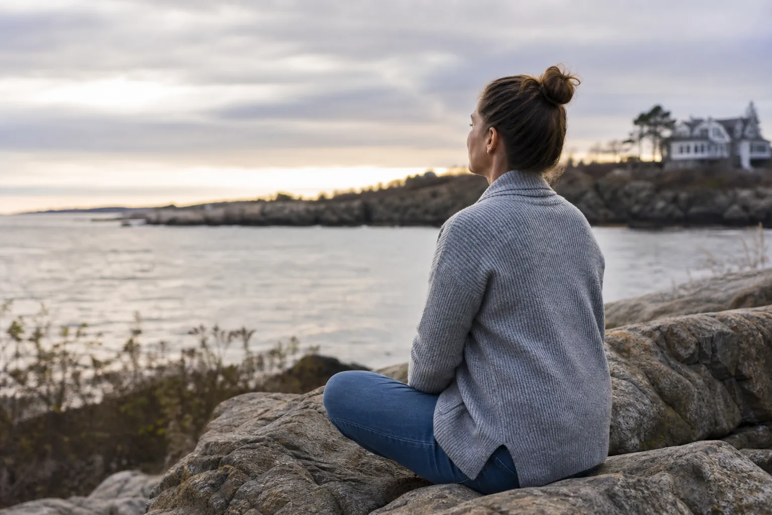 Woman Seated on Coastal Rocks Beside Calm Water A woman in a gray sweater sits quietly on coastal rocks, looking across calm water beneath a cloudy sky.