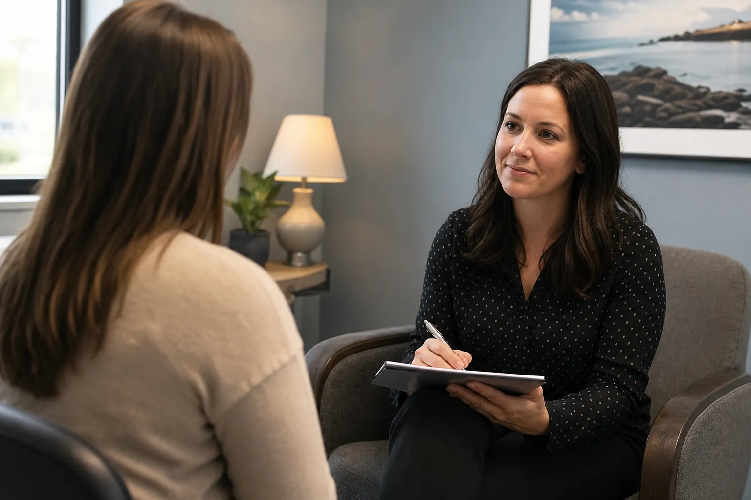 Mental Health Clinic Intake Assessment A mental health clinician holds a notepad while speaking with a patient during an intake assessment in a clinic office.