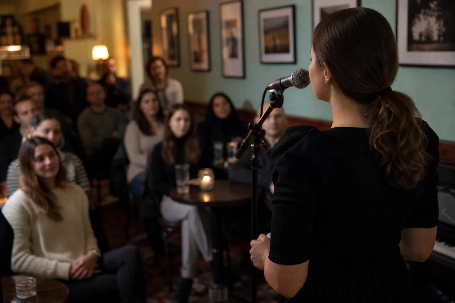 Woman Speaking at a Microphone Before a Small Audience A woman with a ponytail stands at a microphone facing a seated audience in a warmly lit cafe with framed wall art.