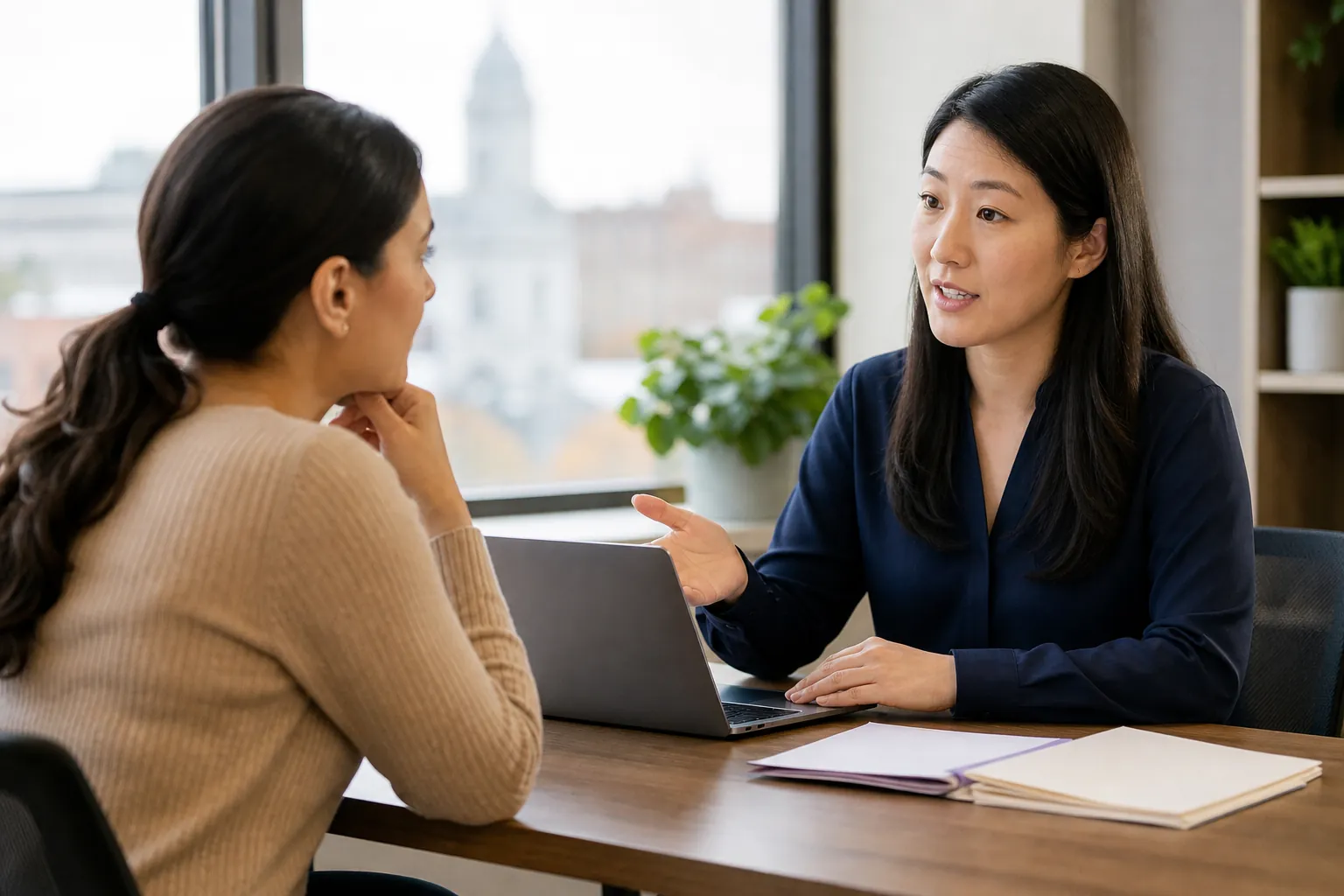 Two women discuss health program coverage across a desk with an open laptop and folders in a bright office.