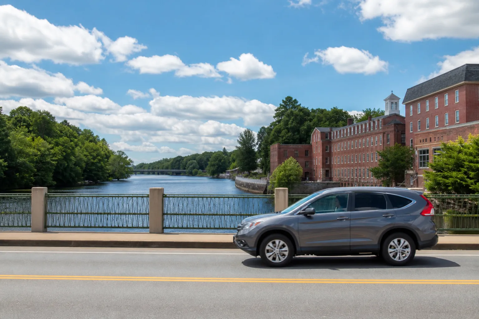 Gray SUV crossing a bridge beside a river and historic red brick mill buildings under a blue sky